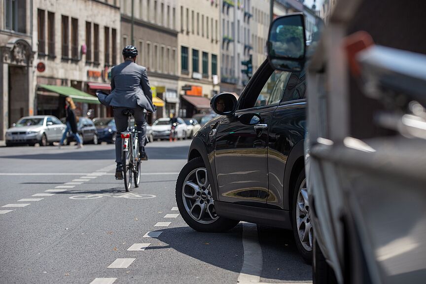 Radverkehr im Stadtverkehr Radfahrer in dunkler Kleidung mit Helm fährt auf einer Stadtstraße, im Vordergrund steht ein schwarzes Auto am Straßenrand, weitere Fahrzeuge und Gebäude im Hintergrund.