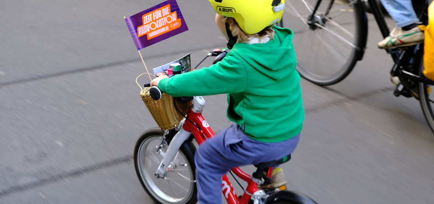 Kidical Mass Friedrichshain/Lichtenberg Ein Kind fährt auf einem roten Rad mit Radvolutionsflagge am Lenker.