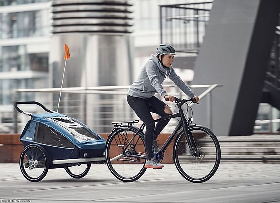 Das Fahren mit Kinderanhänger sollten Eltern üben. Eine Frau fährt auf einem Fahrrad mit einem Kinderanhänger.