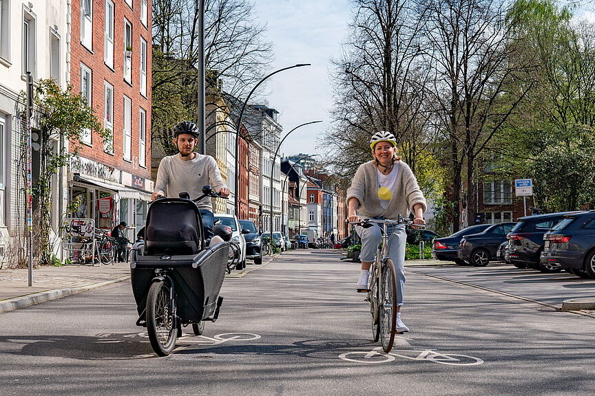 Radfahren auf der Fahrbahn Zwei Personen fahren mit einem Lastenrad mit Transportbox und einem Cityrad auf einer Straße mit Rad-Markierungen; rechts stehen parkende Autos.